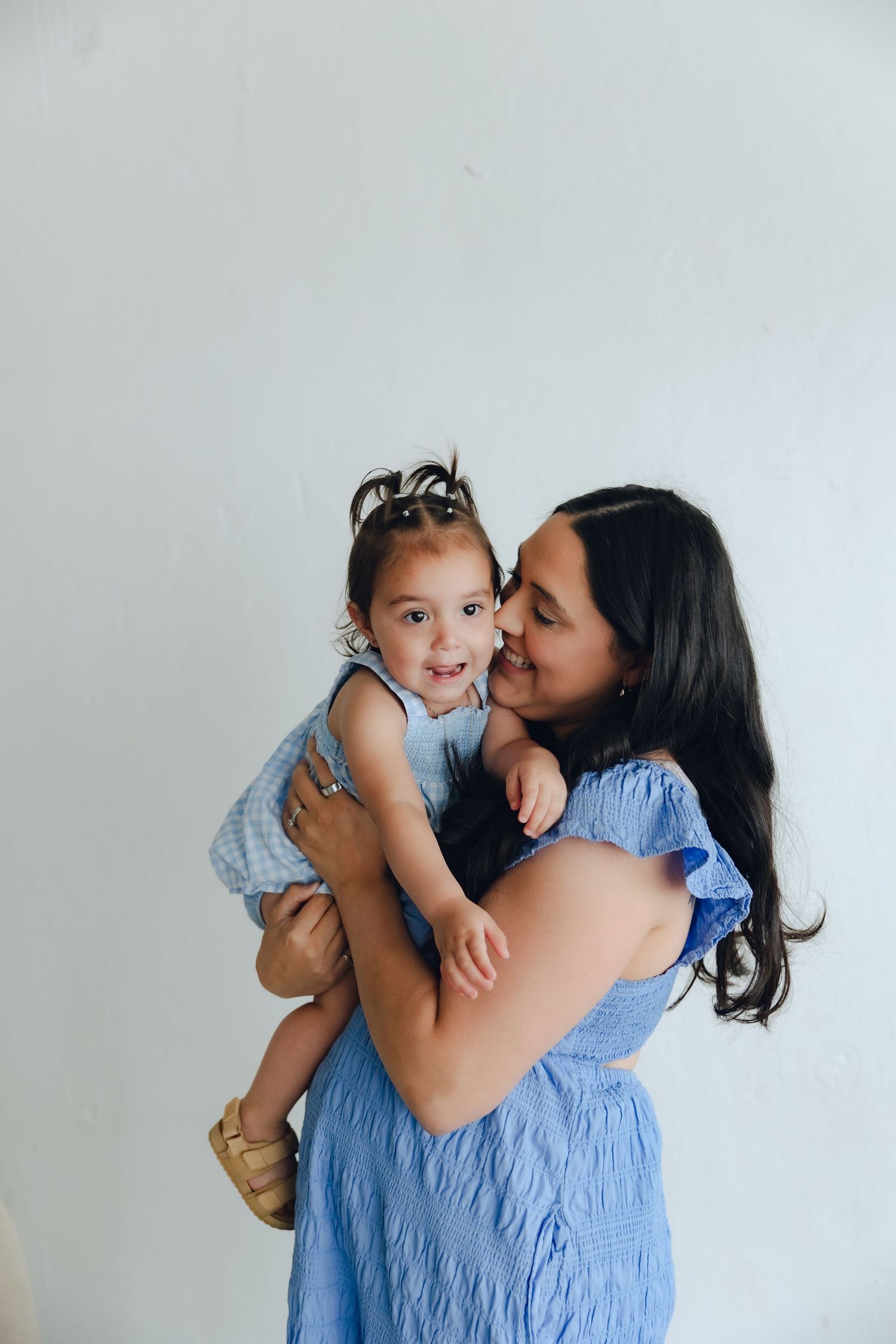 Two people in matching blue dresses share a tender moment against a white wall.