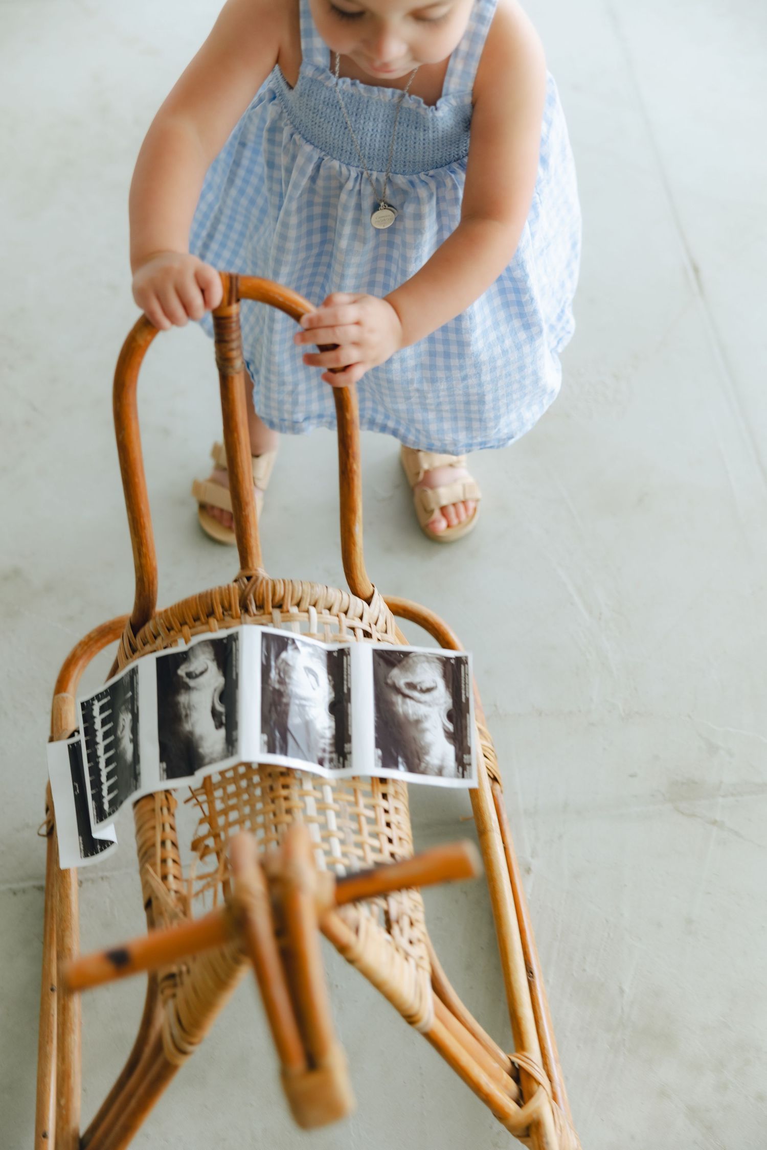 A small child in a blue checkered dress reaches down to touch a wooden rocking chair with photos displayed.