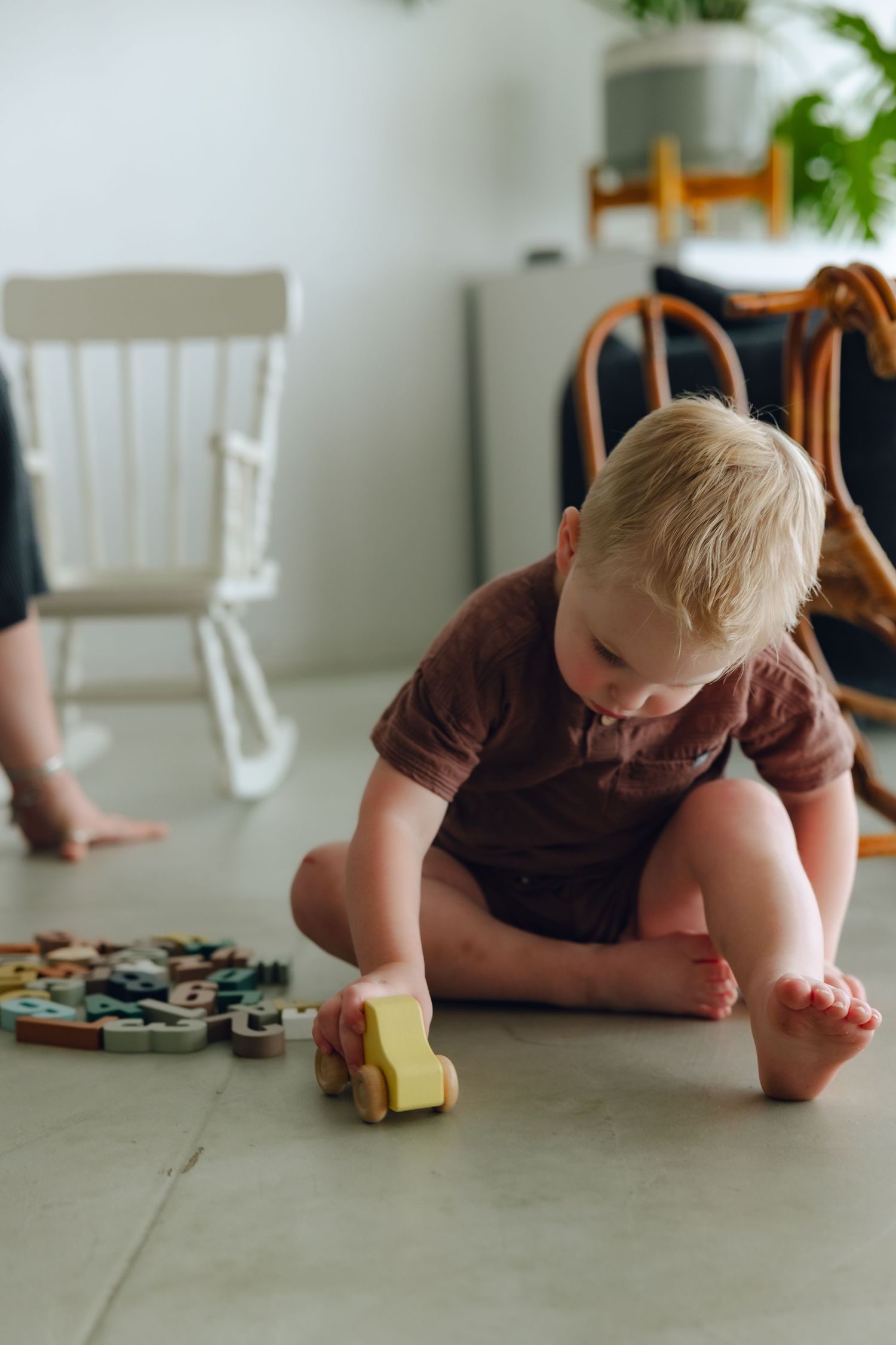 Small child in brown clothing plays with a wooden toy car on a light colored floor while puzzle pieces are scattered nearby.