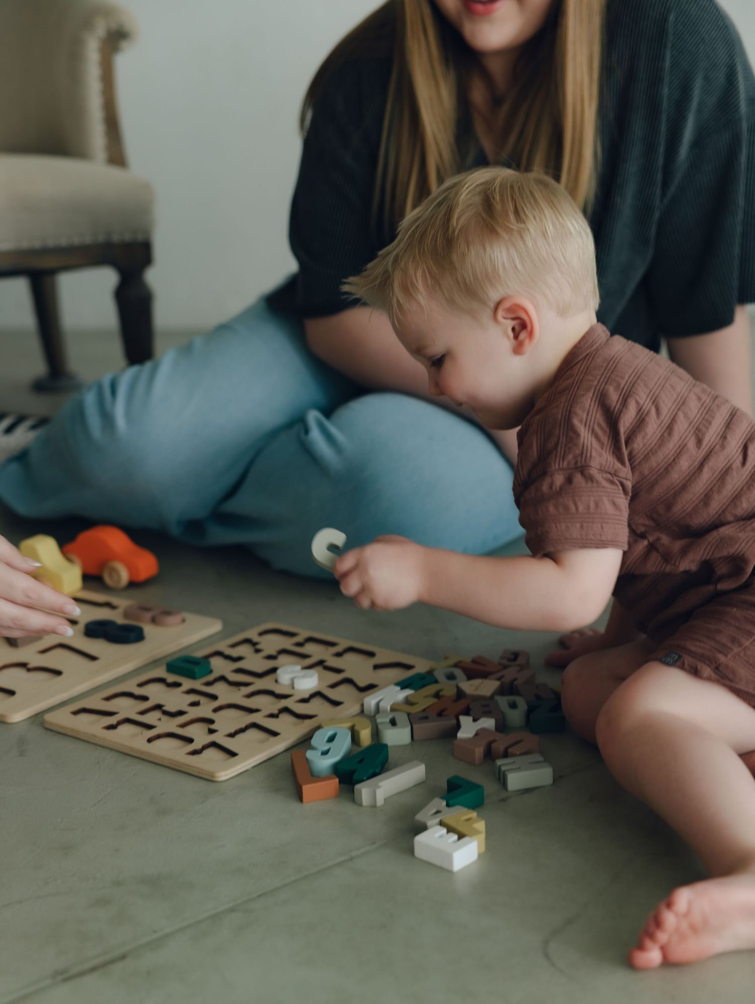 Young toddler in brown outfit plays with wooden puzzle pieces and blocks on floor while someone sits nearby.