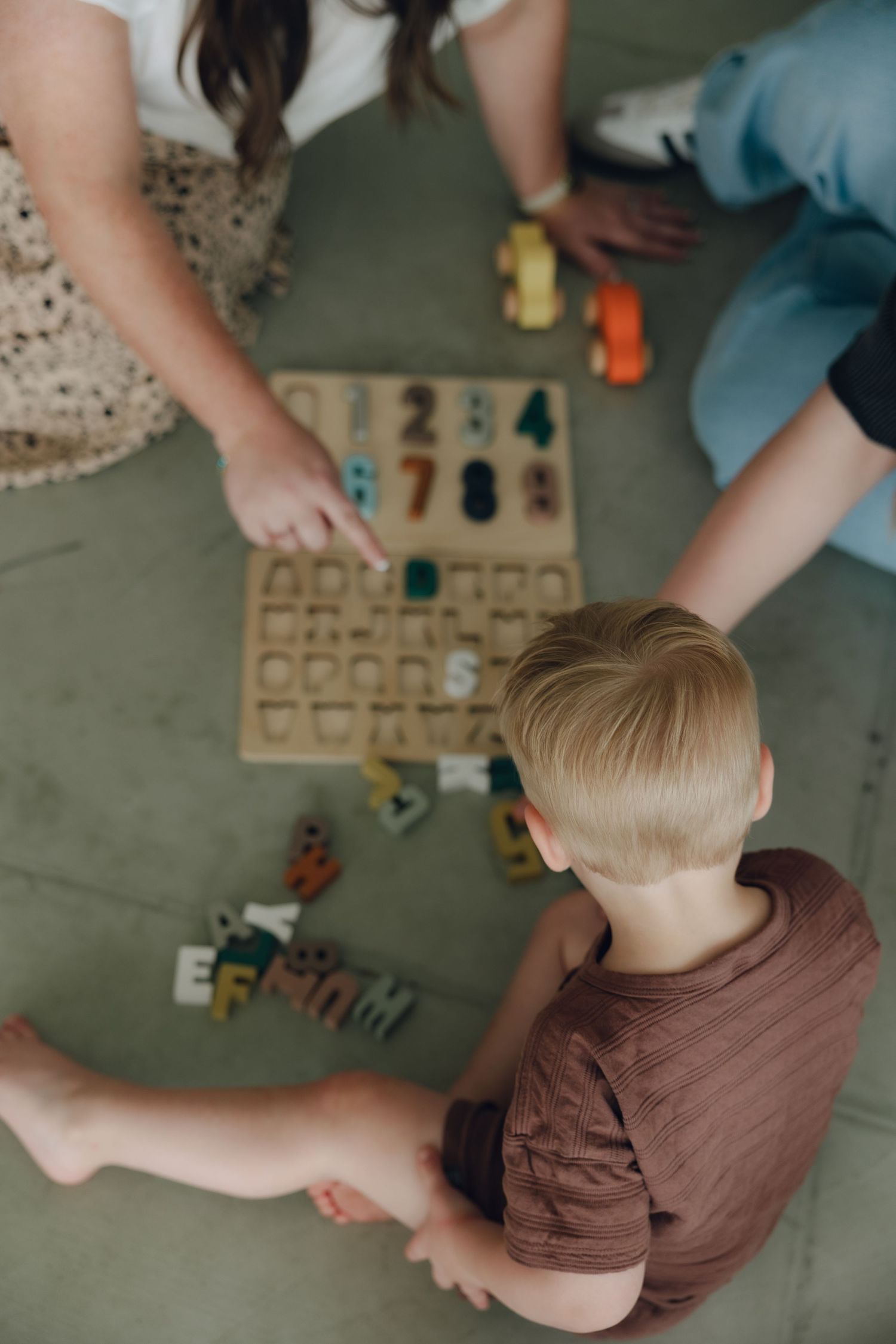 Overhead view of child and adults playing with wooden number blocks and educational toys on floor.