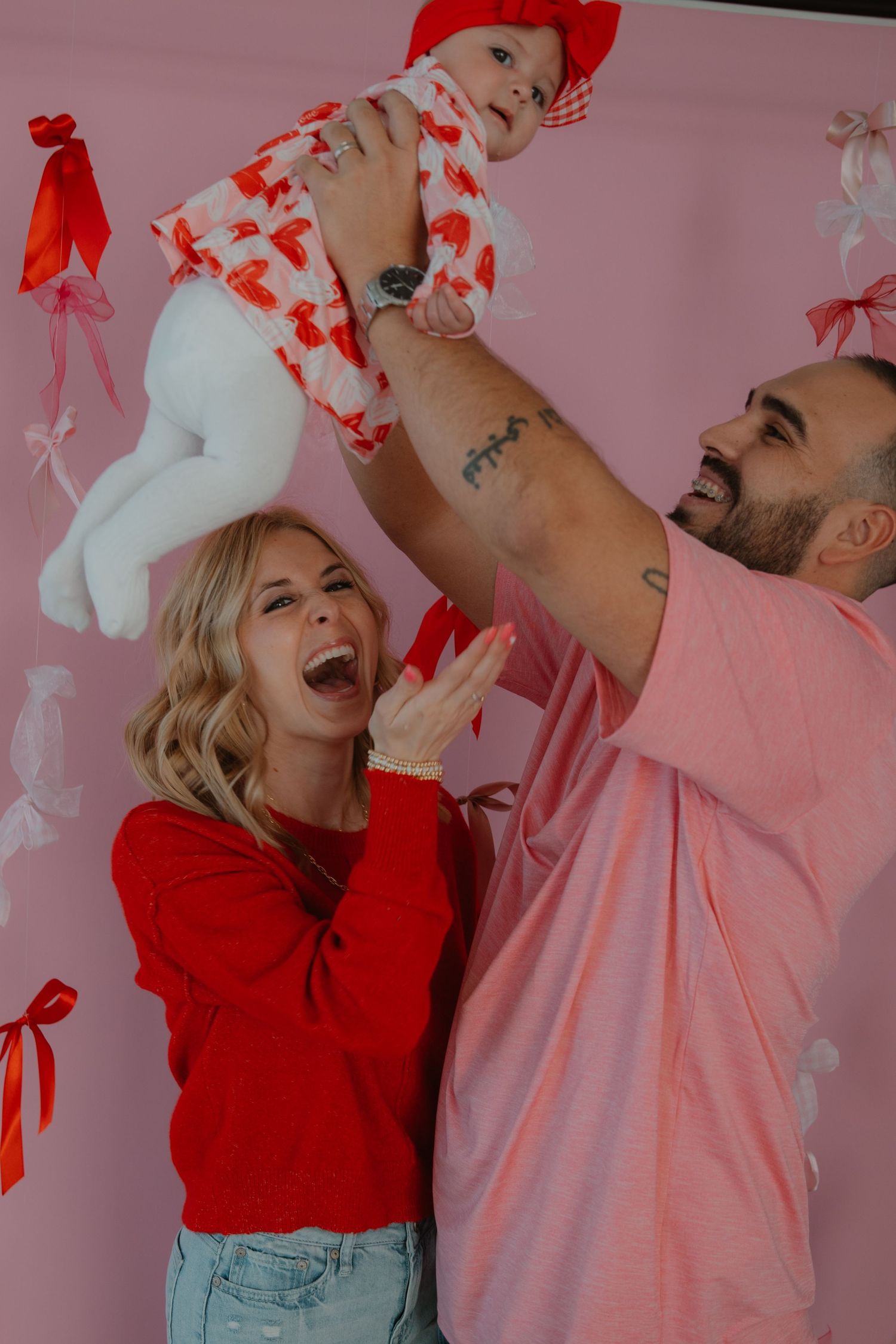 A joyful family moment with a baby in red and white against a pink background.