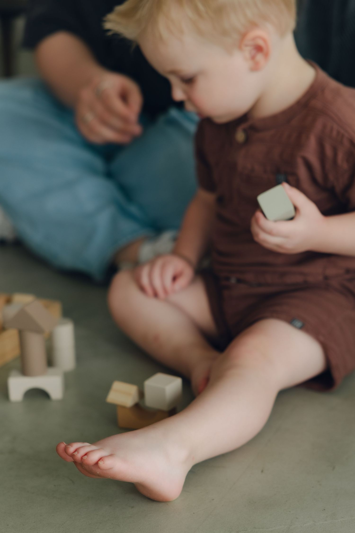 Young toddler in brown outfit plays with wooden blocks while sitting on floor with bare feet.