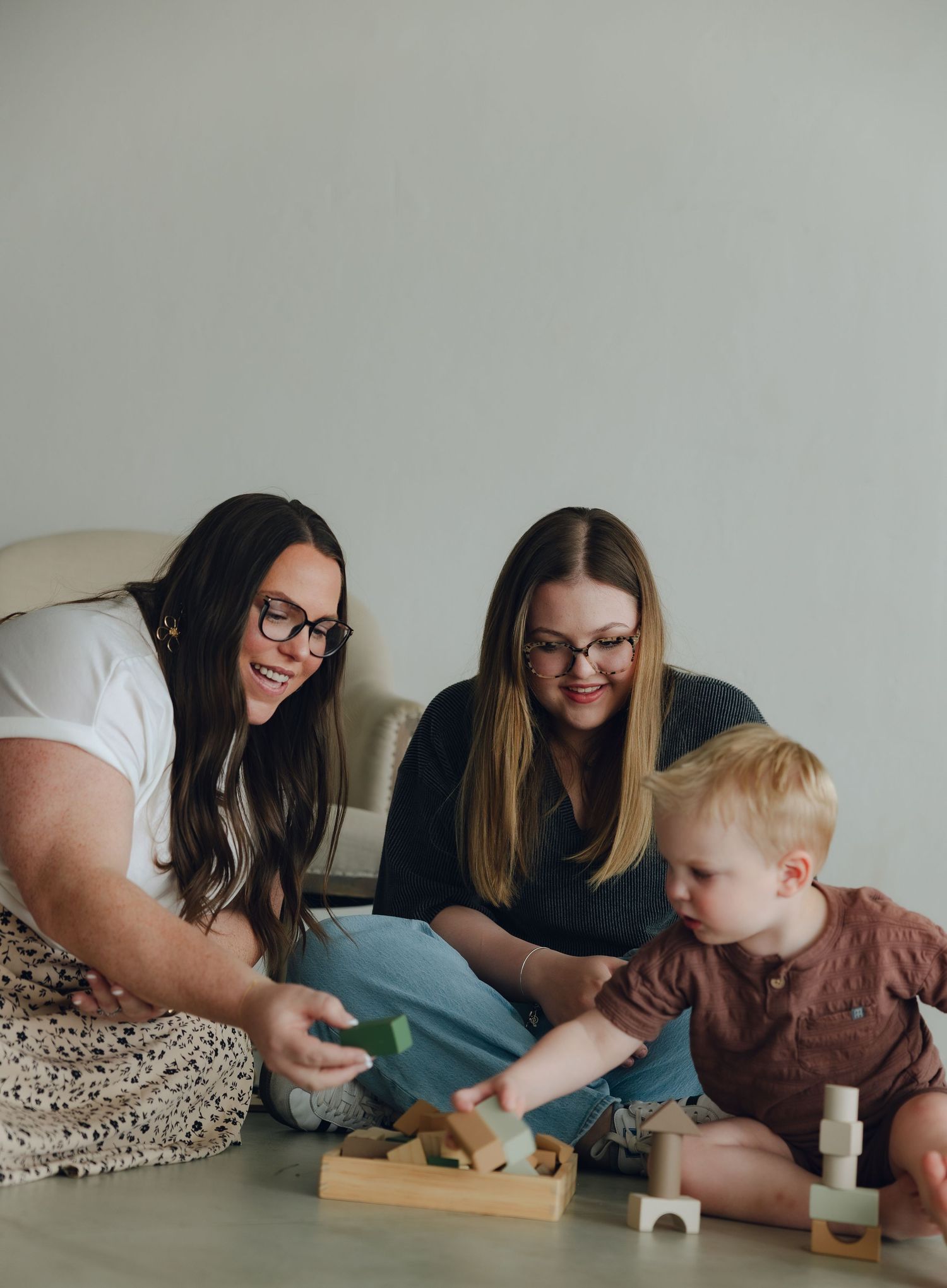 Two adults and a toddler sit on the floor playing with wooden blocks and toys together.