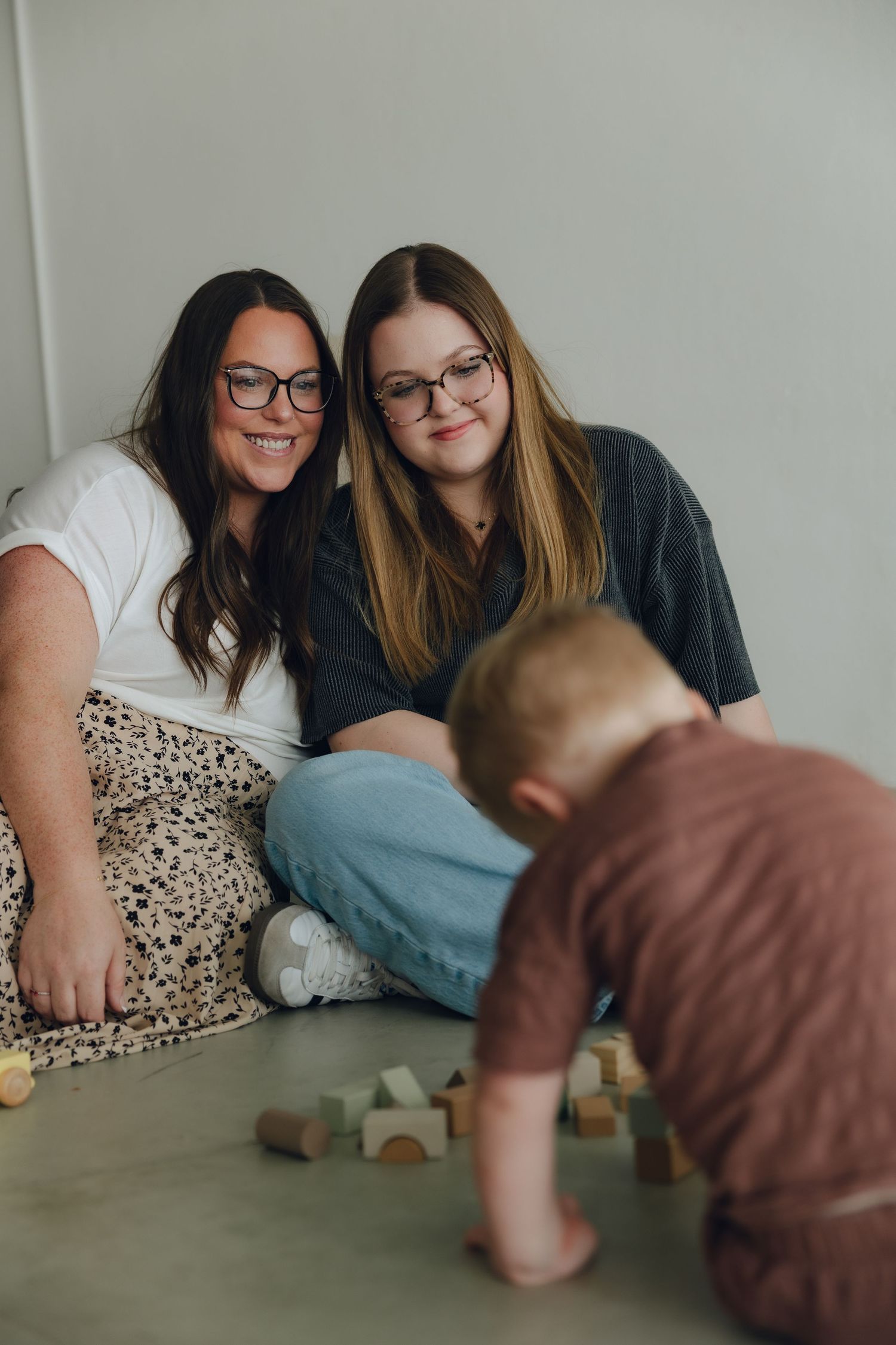 Two people in glasses sitting on floor watching toddler play with wooden blocks in casual home setting.
