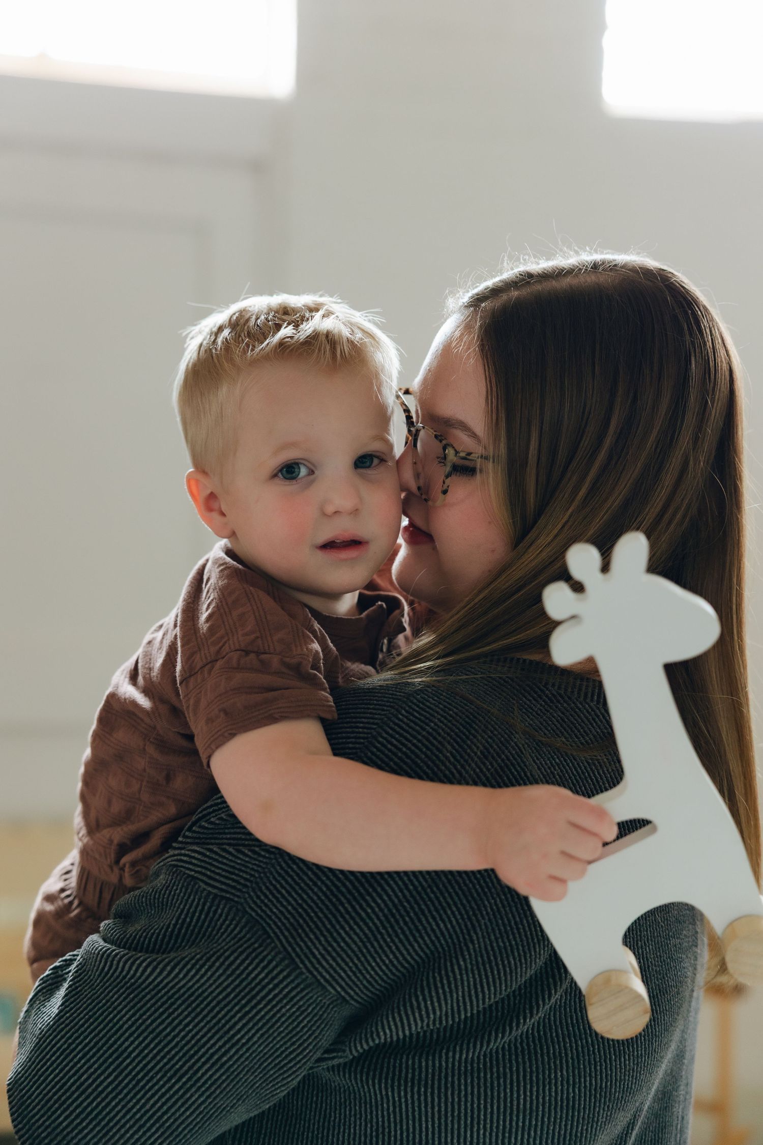 A young child in a brown shirt is being held while holding a white giraffe toy in a sunlit room.