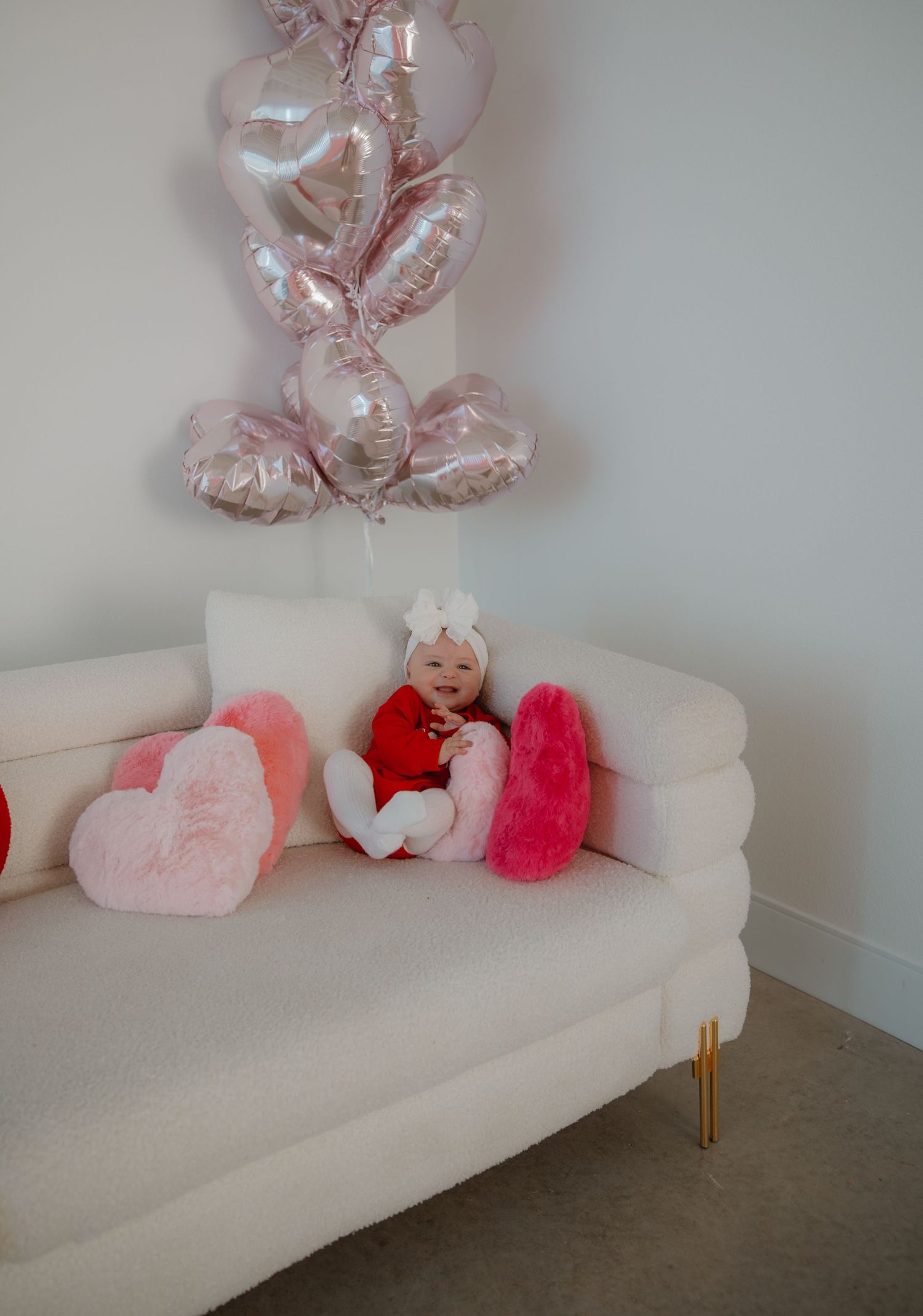 A baby rests on a white couch under pink heart balloons and Valentine's Day decorations.