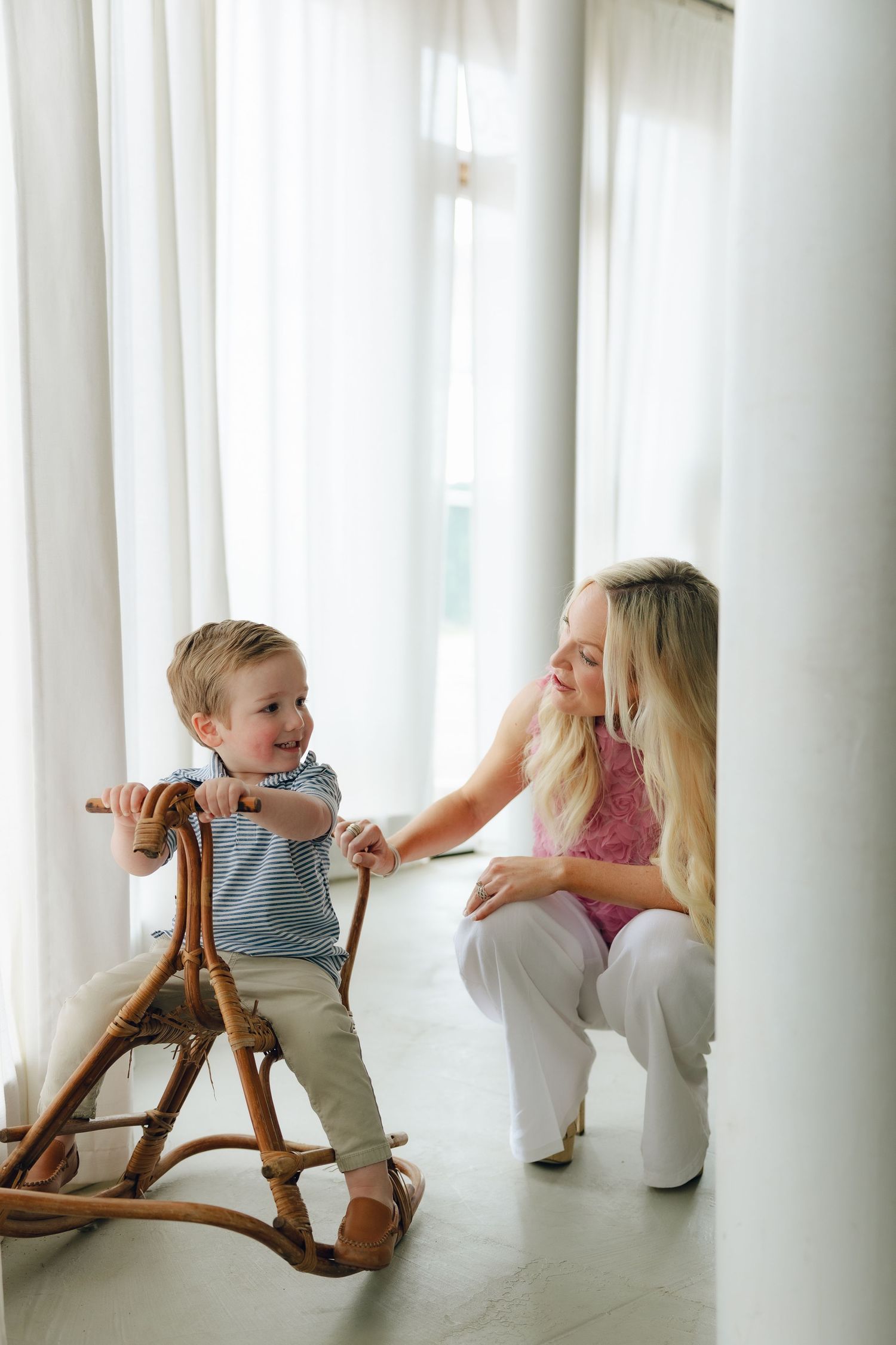 A small child plays on a wooden rocking horse near sheer white curtains in a bright, minimalist room.