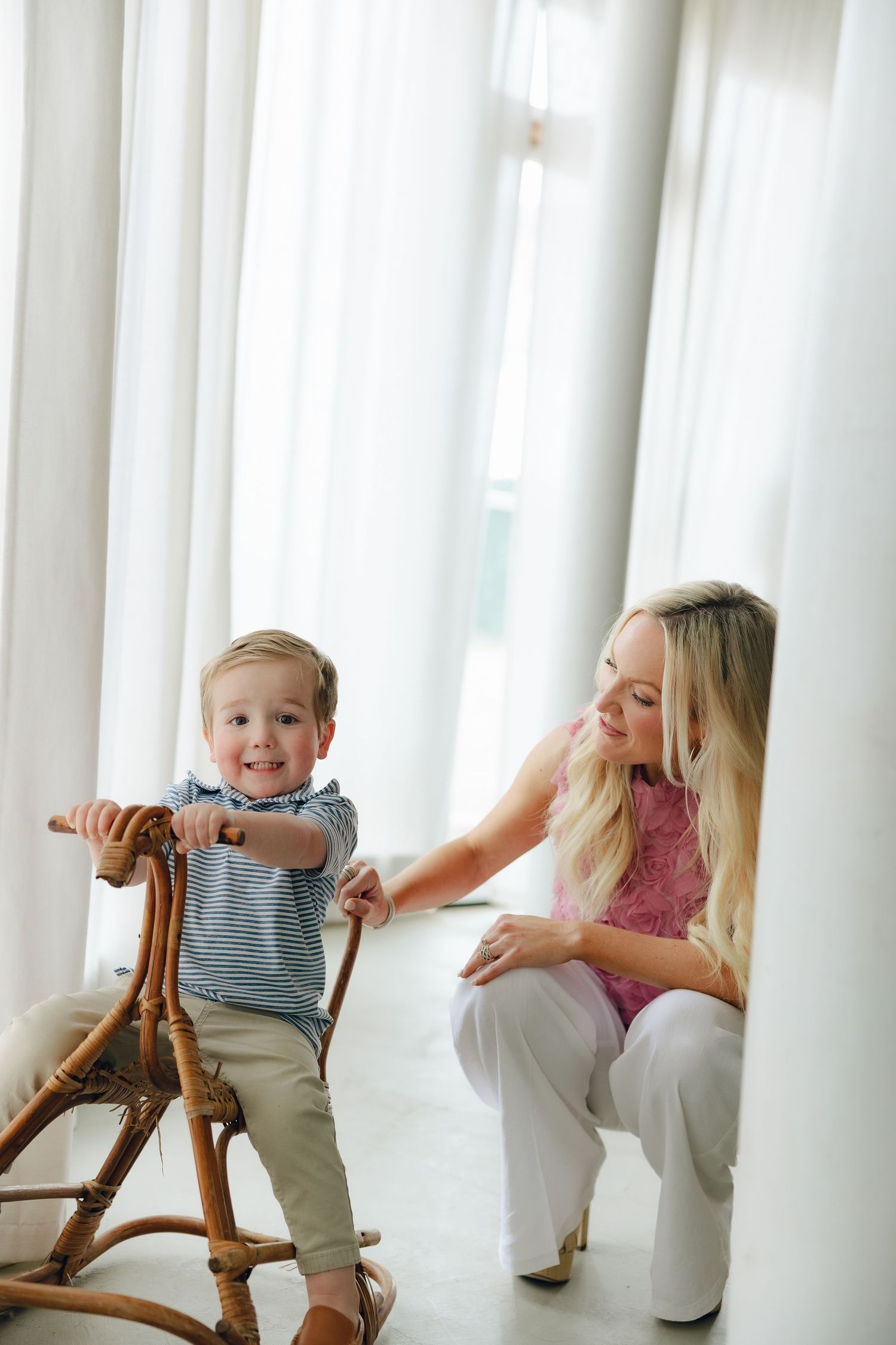 A child enjoys playing on a wooden rocking horse while someone watches nearby in a bright sunlit room.