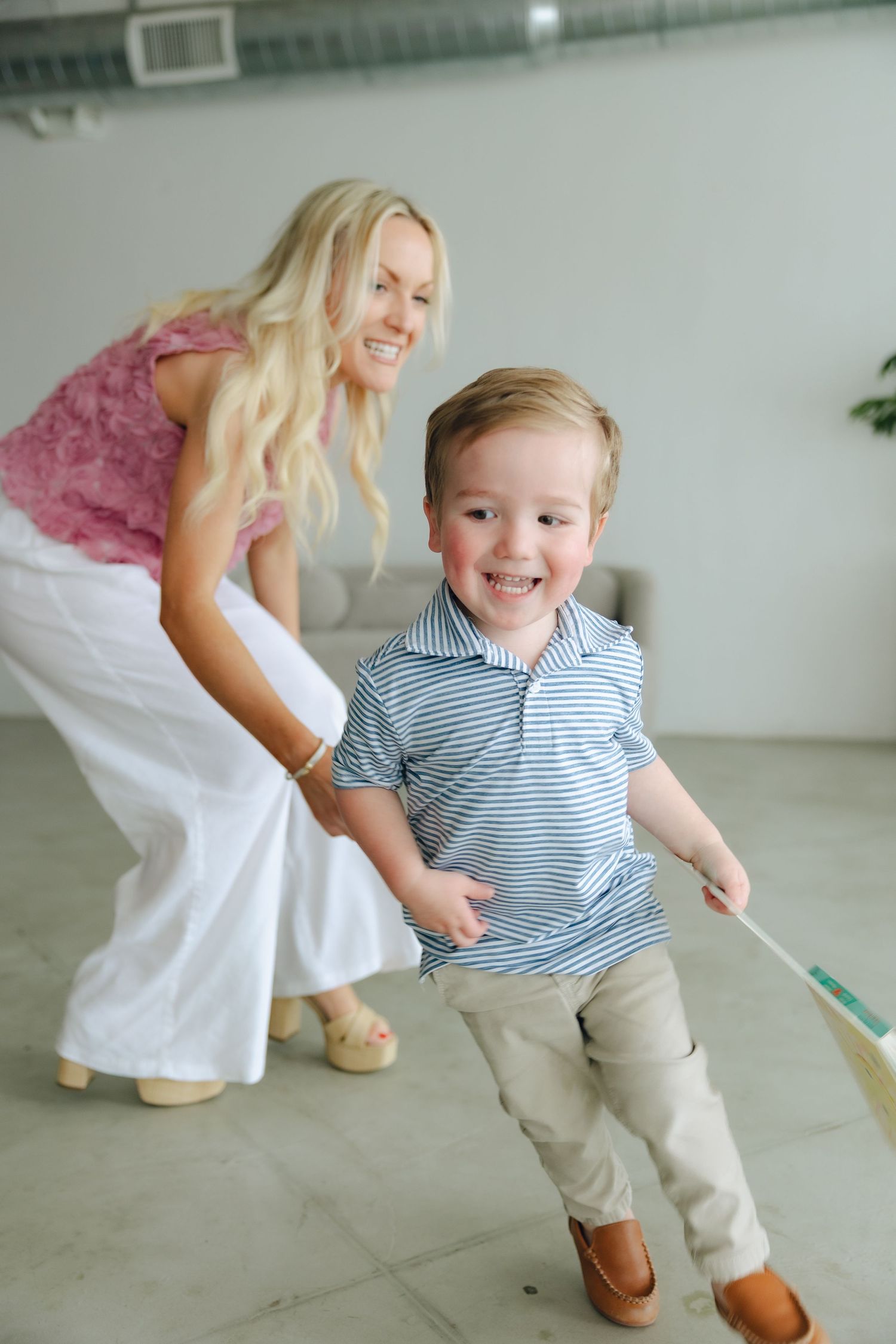 A child in a striped polo shirt and khakis plays indoors while an adult in white pants and pink top leans forward playfully.