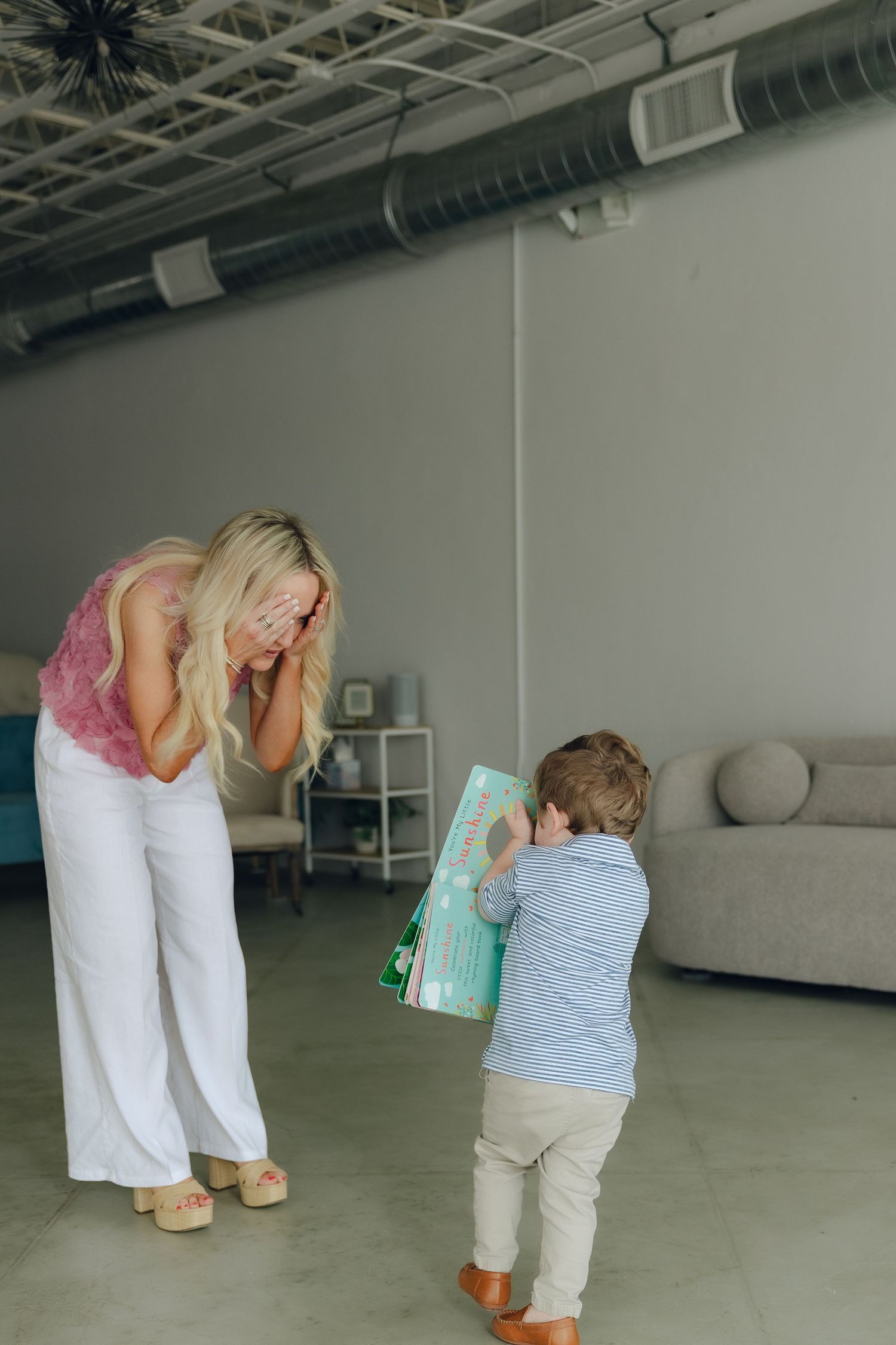 A mother and child share a playful moment in a modern industrial space with exposed ductwork and a gray couch.