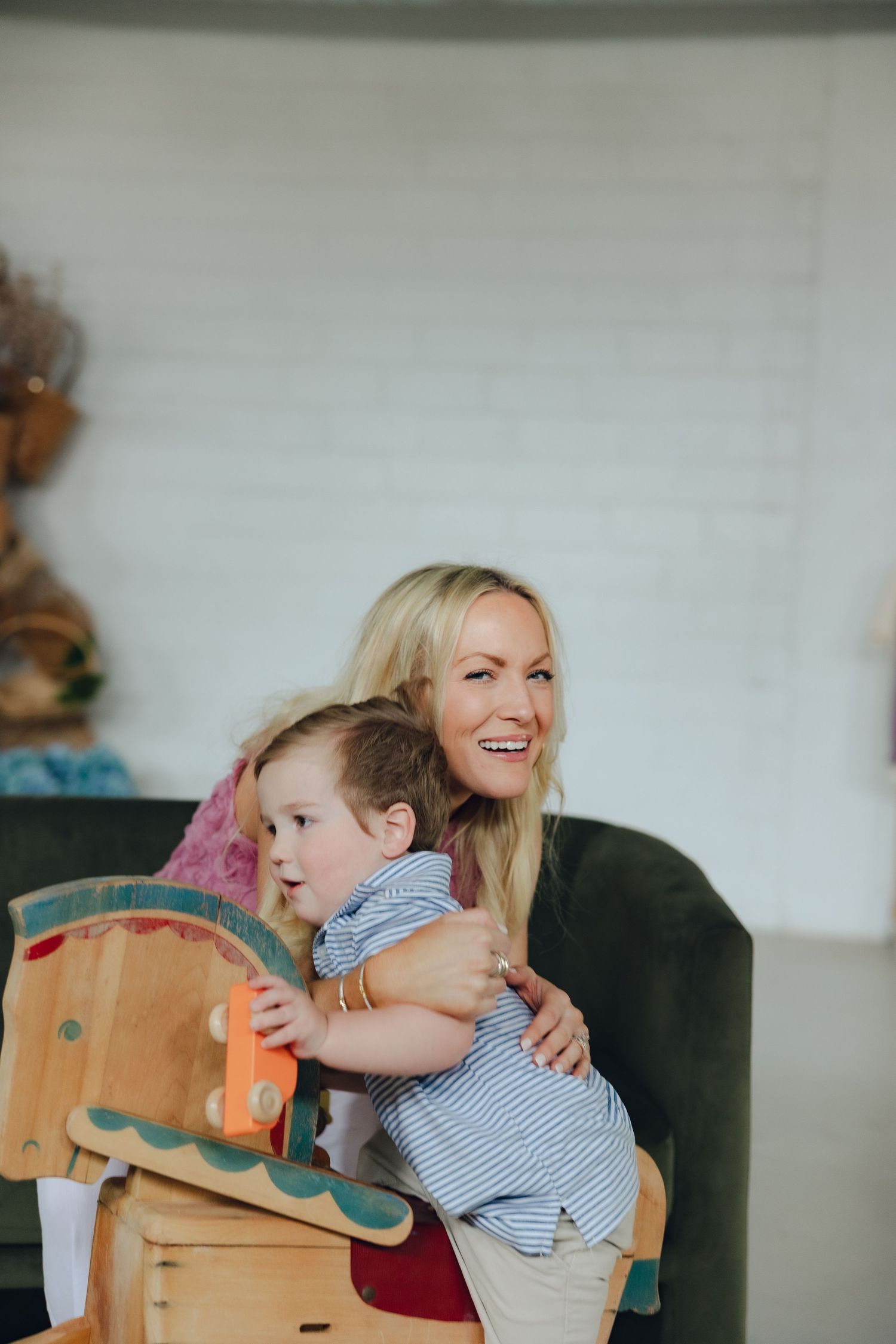 A parent and child share a joyful moment while playing on a wooden rocking horse against a white wall.