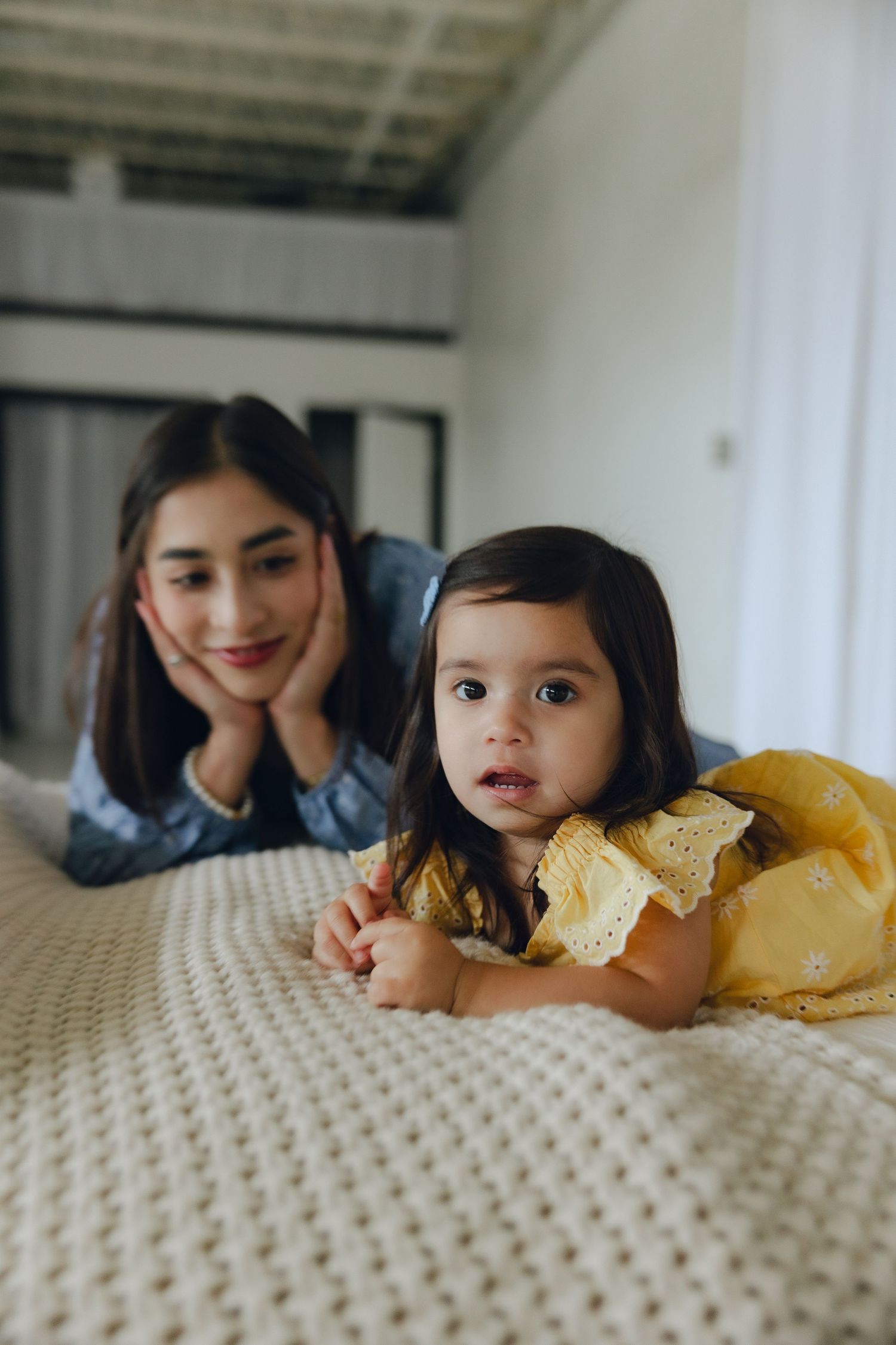 A toddler in a yellow dress lies on a knitted blanket while someone watches from behind.