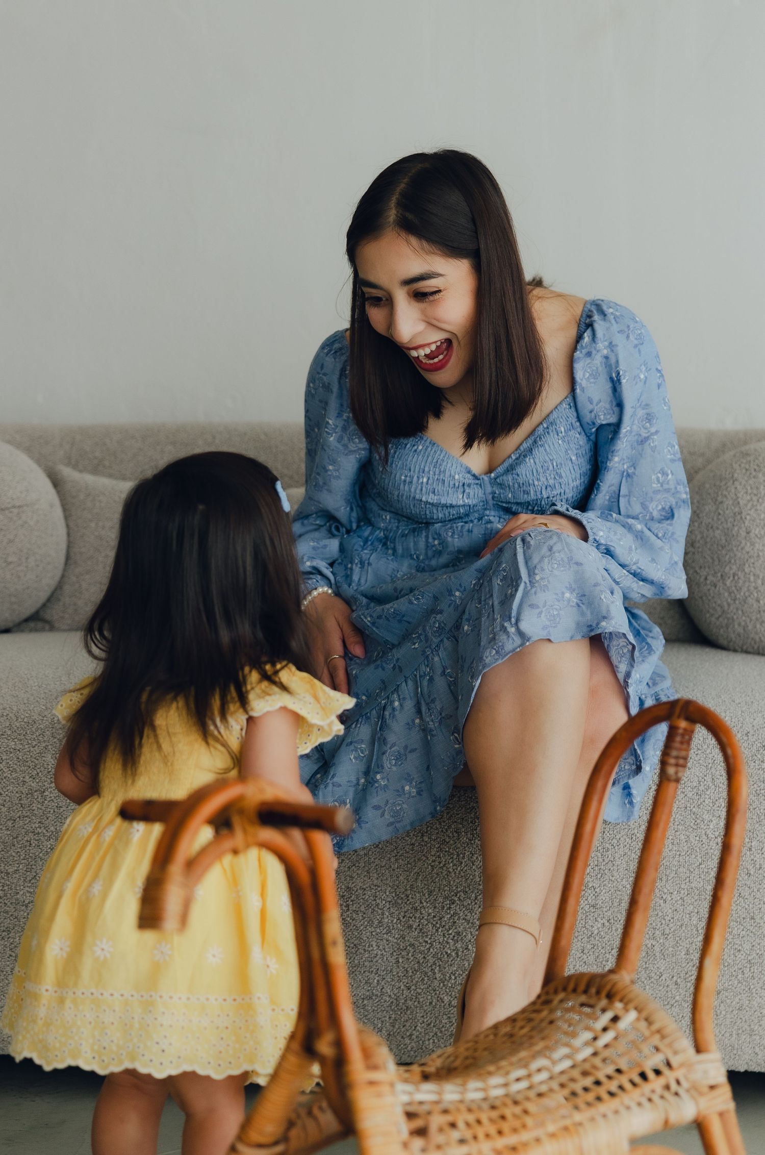 Candid moment shows parent and young child in yellow dress sharing a joyful conversation on a gray couch with wicker bag nearby.