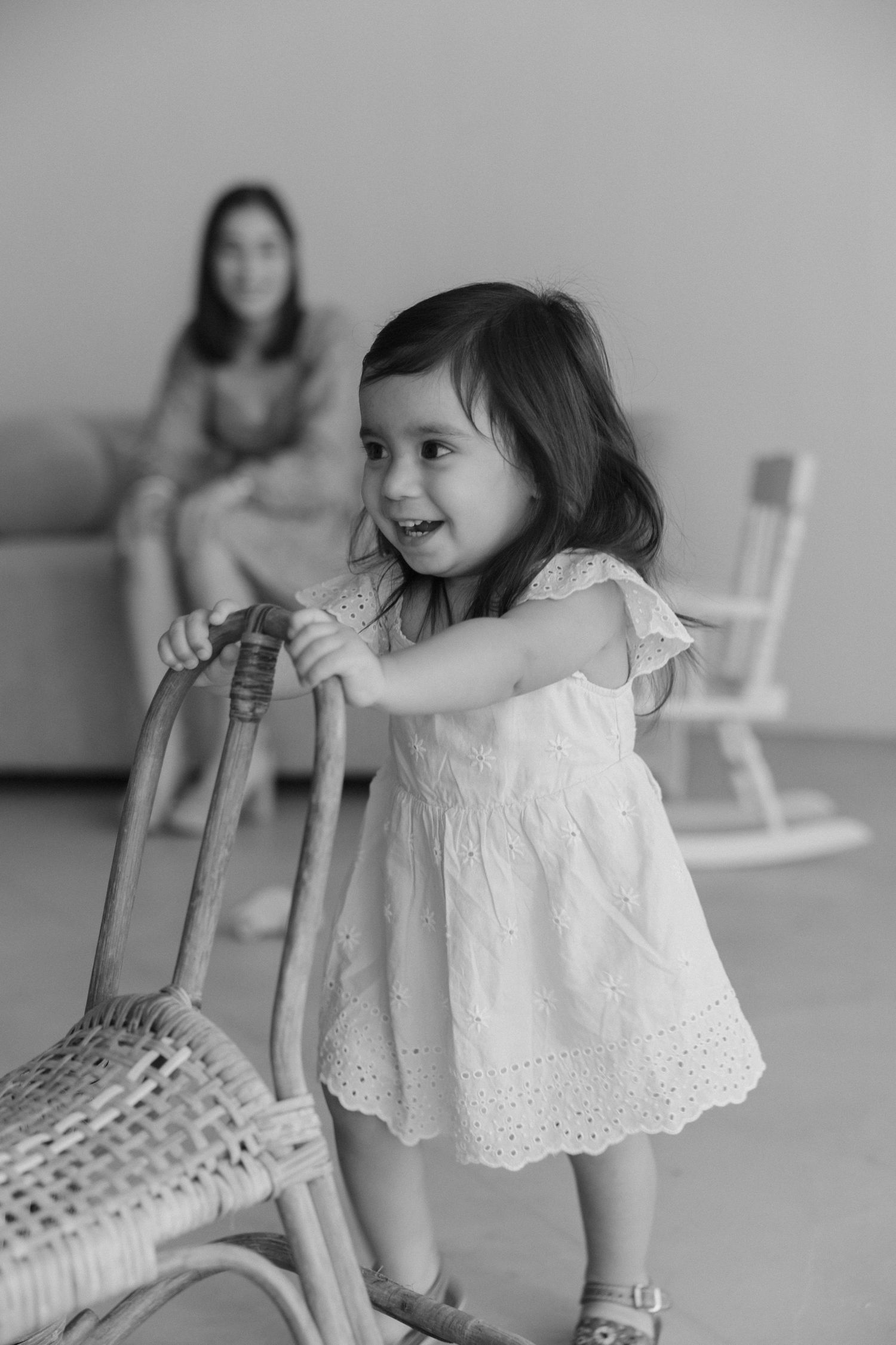 A child in a white eyelet dress plays with a rattan chair indoors while someone sits on a couch in the background.