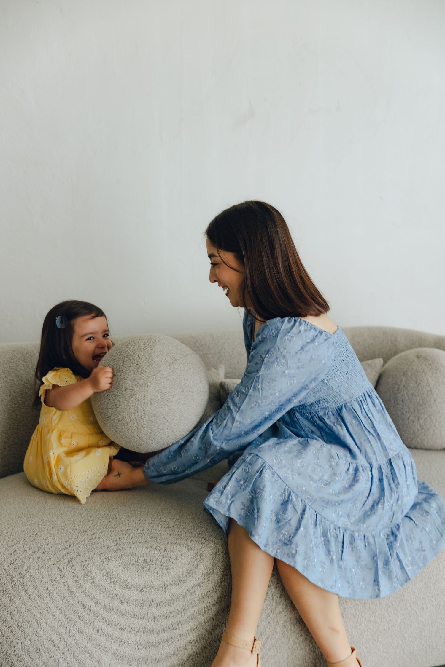 Two people sit together on a gray couch sharing a joyful moment, one wearing a yellow dress and one in light blue denim.