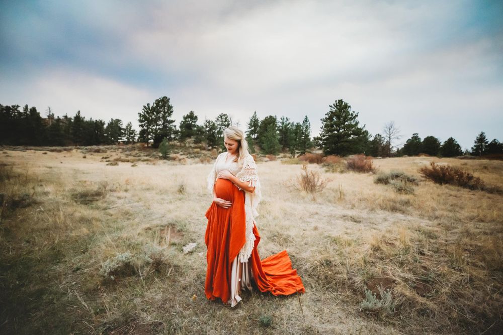 Pregnant woman in white and orange gown holding her belly with trees and field in the background