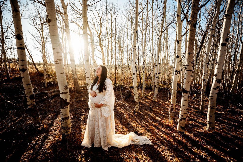 Pregnant woman in white gown holding her belly surrounded by white birch trees at sunset in Dickson, TN