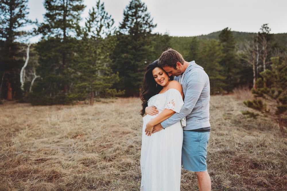 Pregnant woman in white dress with her husband hugging her from behind with mountains in the background