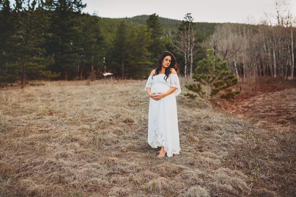 Pregnant woman in white dress holding her belly and smiling in the mountains