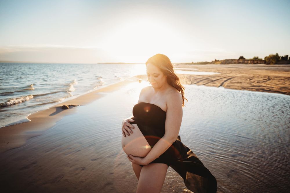 Pregnant woman in black maternity dress holding her belly with beach in the background