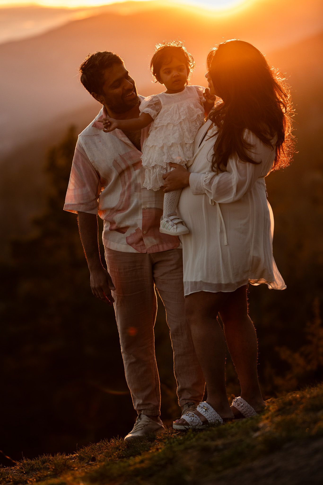 A family stands together on a hilltop silhouetted against a warm golden sunset in the mountains.