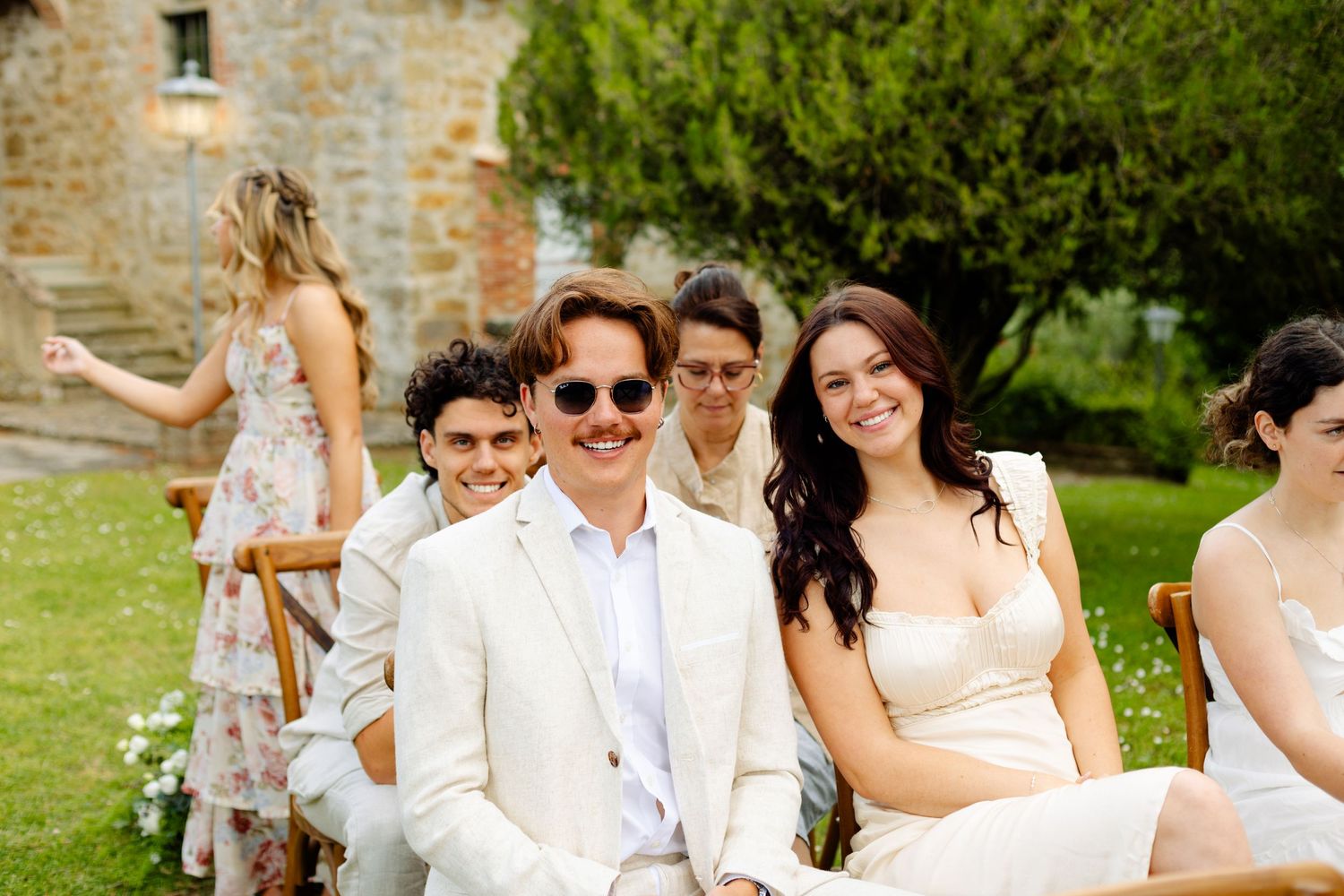 Wedding guests seated outdoors at a rustic stone venue with summer garden scenery.