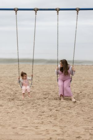 A mother and young daughter sit on swings on the beach.