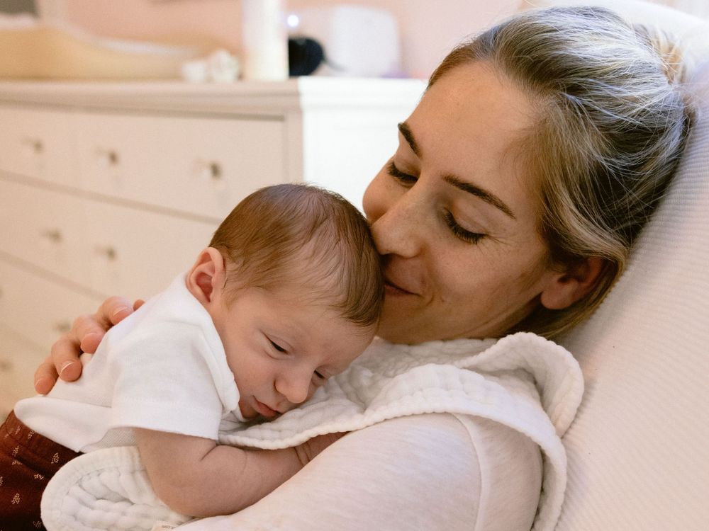 Close-up portrait of a mother smiling and holding a newborn while seated in a soft pink nursery rocking chair.