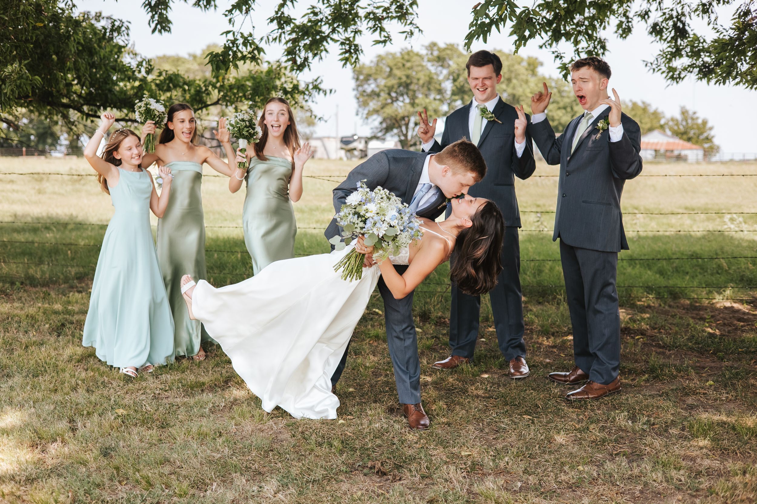 Wedding party poses for outdoor photos on grassy lawn with trees in background.