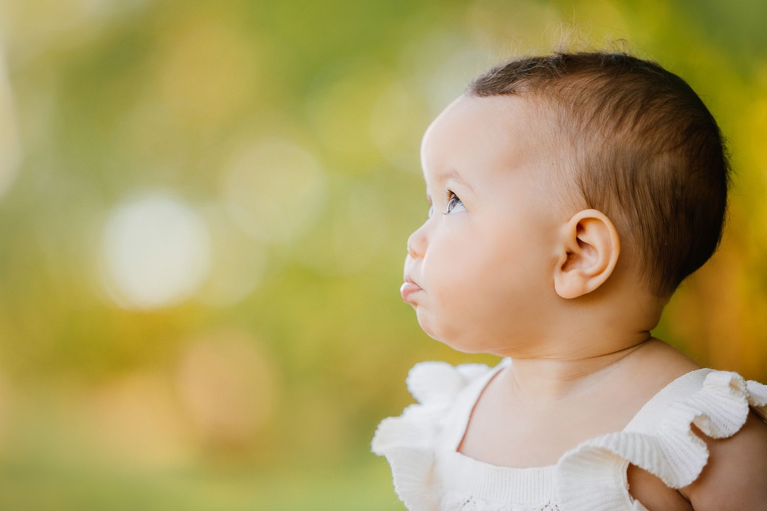 Baby in white dress looking to side against blurred yellow-green background.