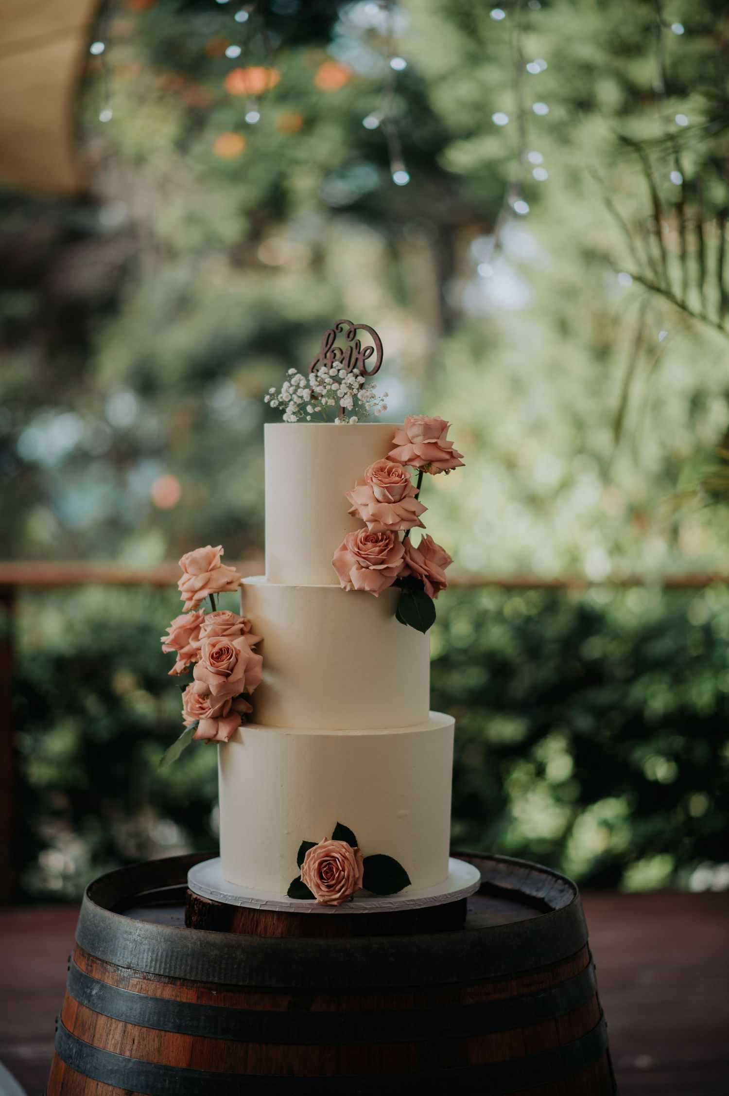 Elegant three-tier white wedding cake with peach roses displayed on rustic wine barrel.