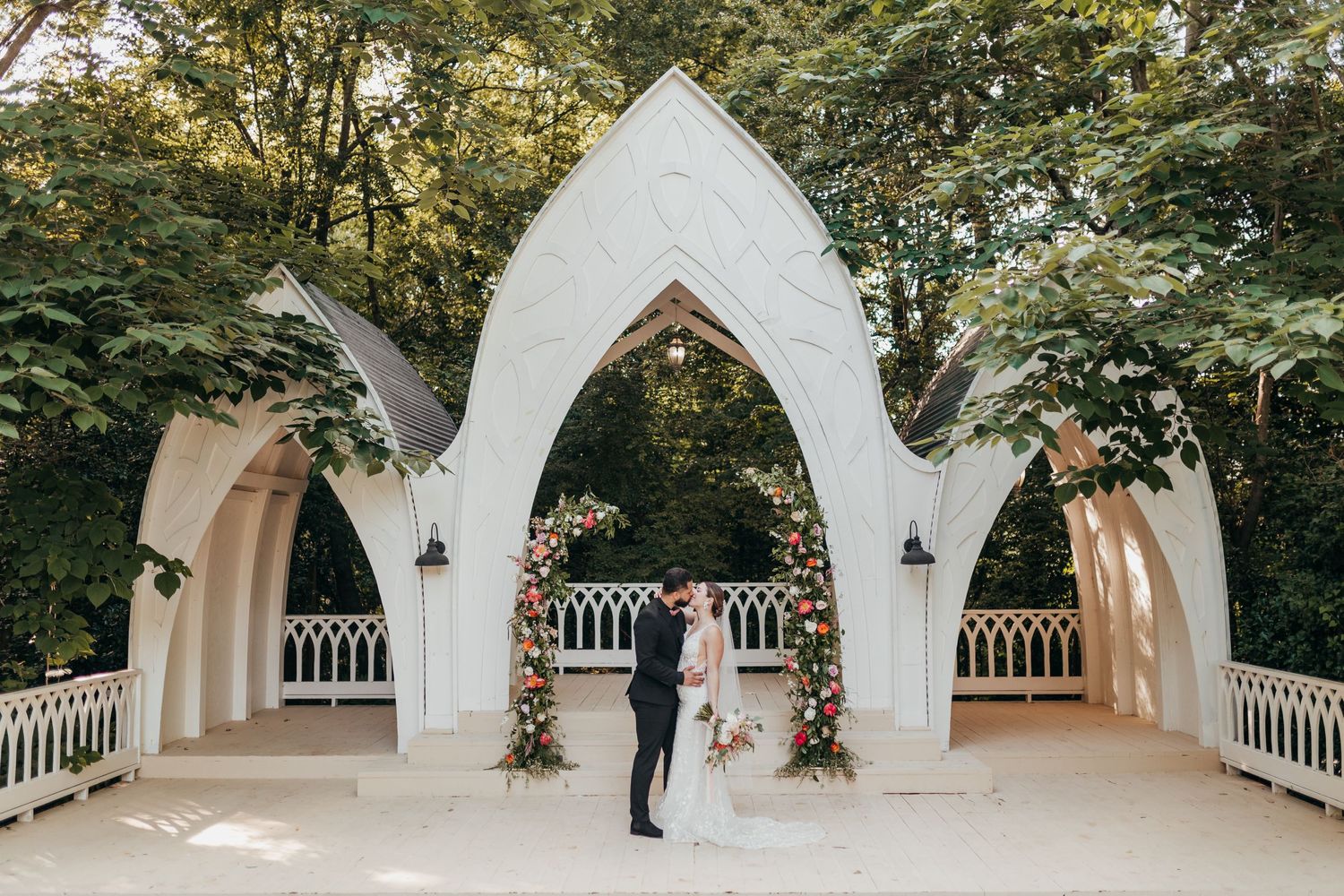 Wedding ceremony under white gothic arch decorated with floral arrangements.