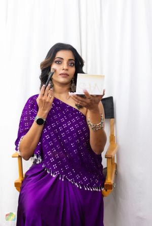 A person in a purple sequined sari applies makeup while sitting in a chair.