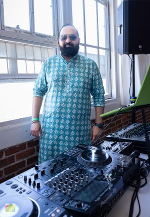 DJ in turquoise patterned kurta stands behind professional mixing equipment.