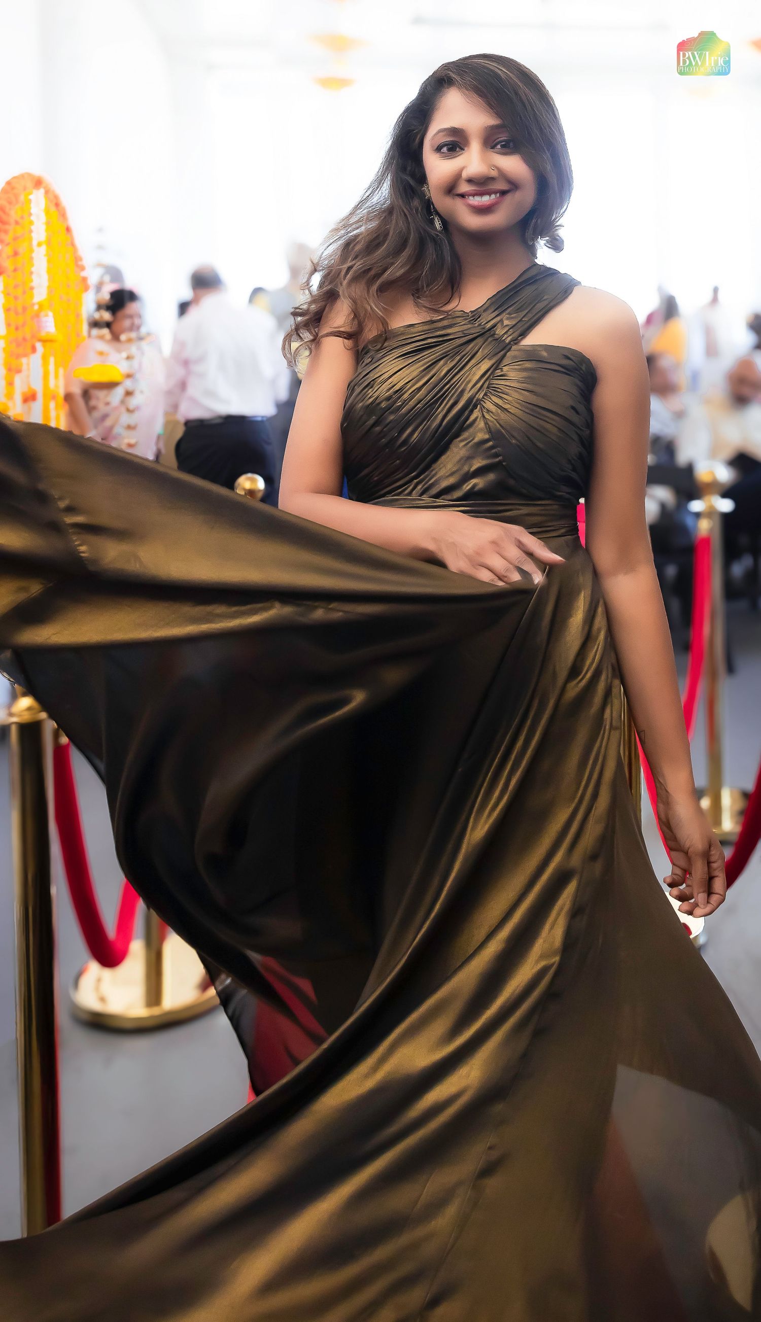 Model poses in flowing metallic gold evening gown on red carpet between velvet ropes and stanchions.
