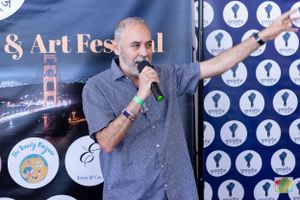 A speaker gestures while holding a microphone at an arts festival with San Francisco backdrop.