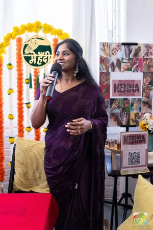 A speaker in purple attire presents at an event with yellow marigold decorations and promotional banners.