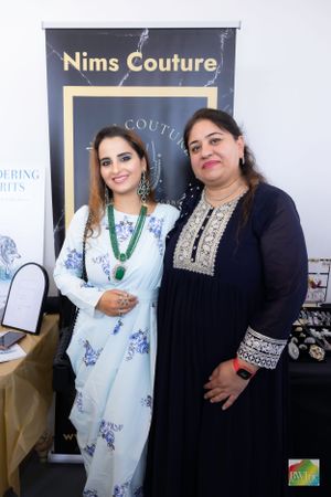 Two women pose next to a Nims Couture branded banner at a fashion event.