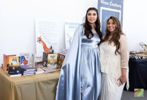 Two women in formal wear pose at promotional event with display items on table.