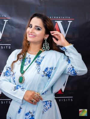 A woman in a light blue floral dress with ornate jewelry poses against a dark backdrop.