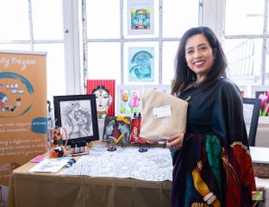 Artist showcasing artwork and merchandise at creative vendor booth with window lighting.
