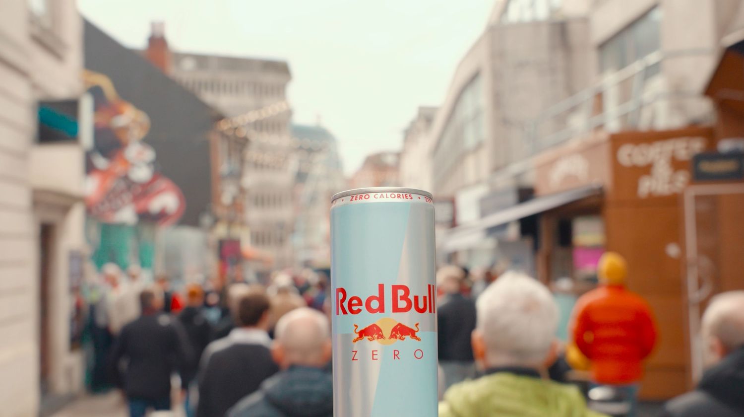 A can of Red Bull energy drink held up against a blurry urban street background with people walking.