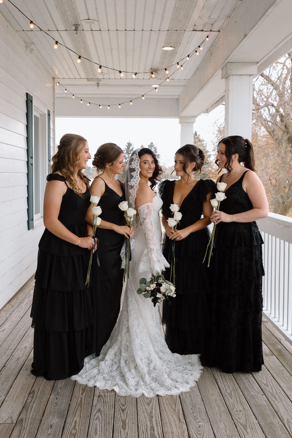 Bridal party in black dresses and white gown posing on a covered porch with string lights.