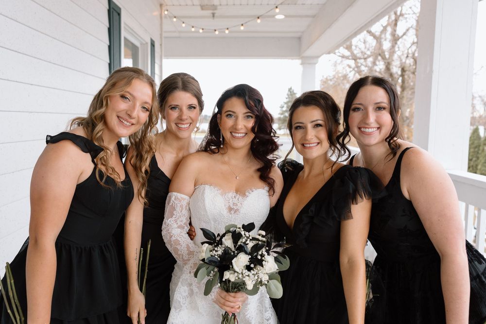 Wedding party lined up on white porch wearing black dresses and holding bouquets.