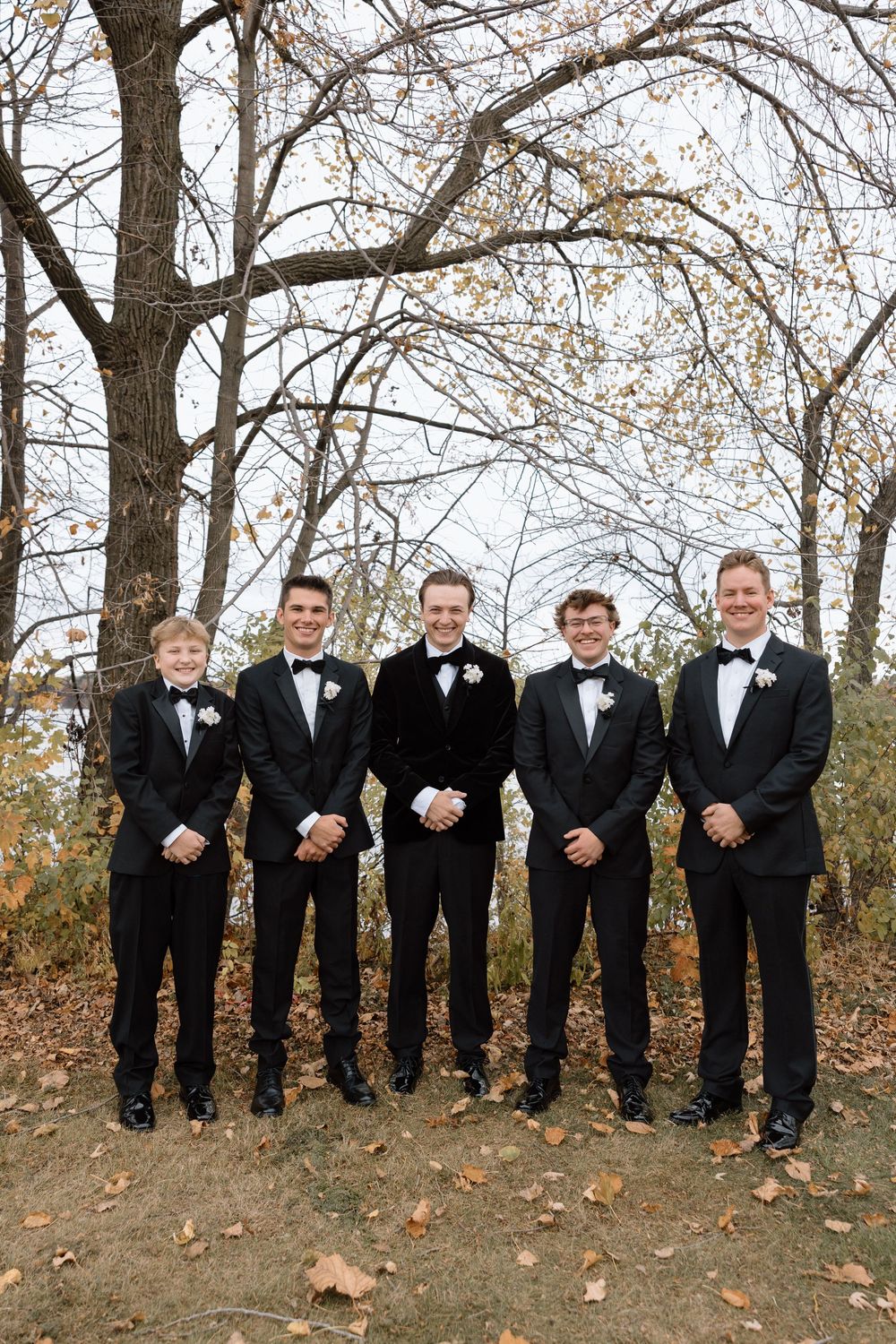 Groomsmen in black tuxedos standing in line against autumn trees.