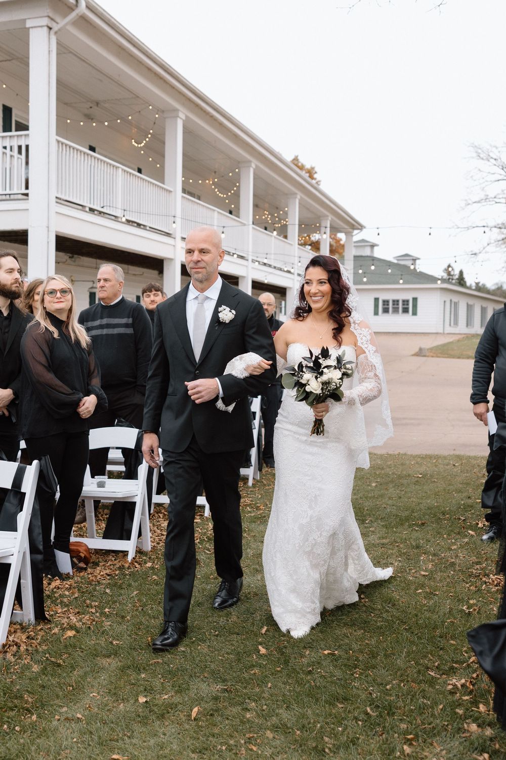 Wedding processional walking outside white columned building with autumn leaves.