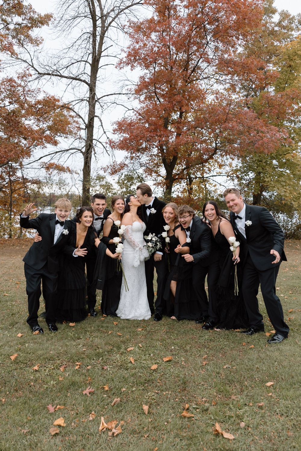 Large wedding party in black attire posing together on grassy hill during autumn.