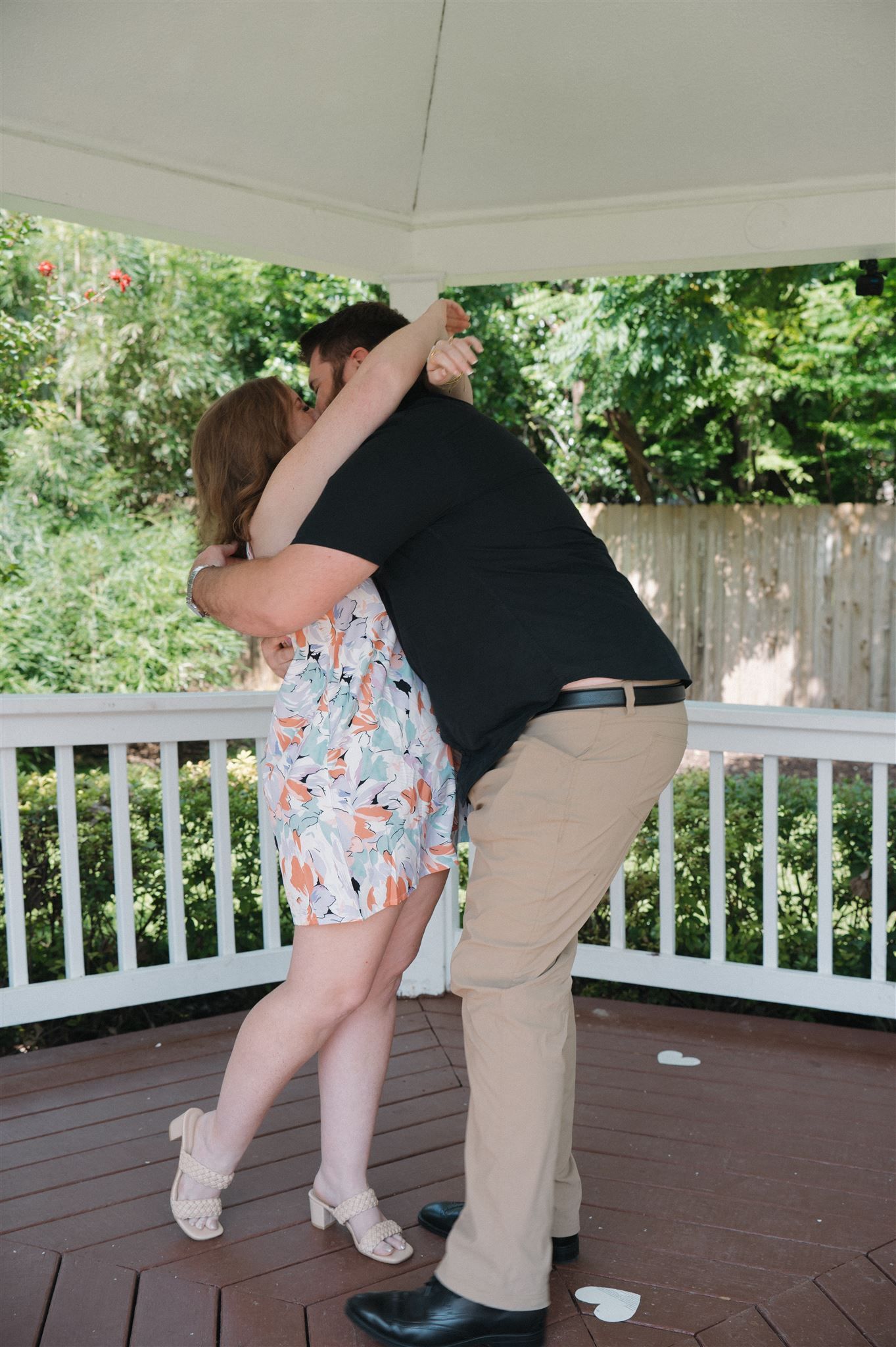A couple embraces and shares a romantic moment on a wooden deck with white railings surrounded by green foliage.