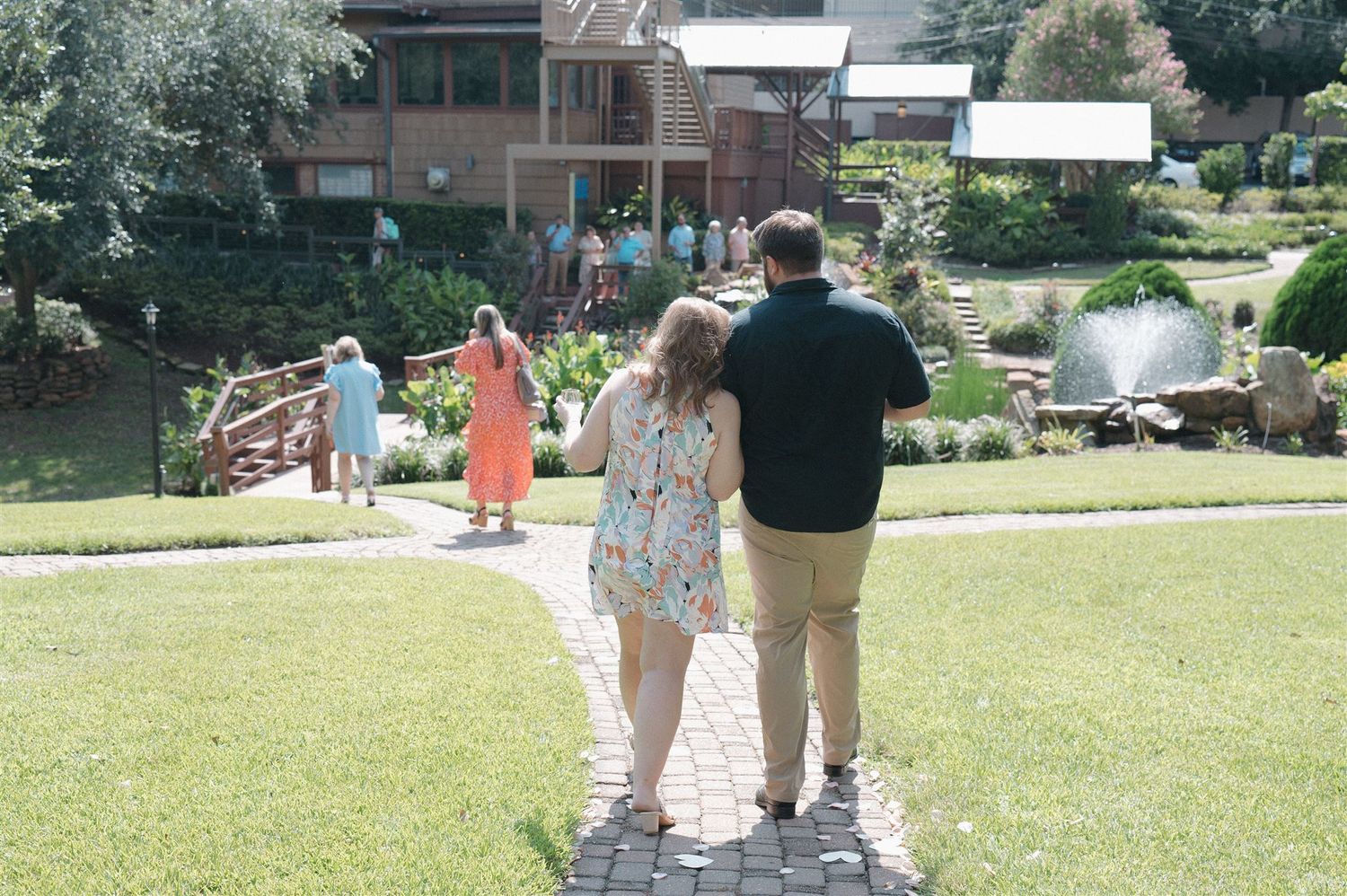 A couple walks along a stone path through a lush green garden on a sunny summer day.
